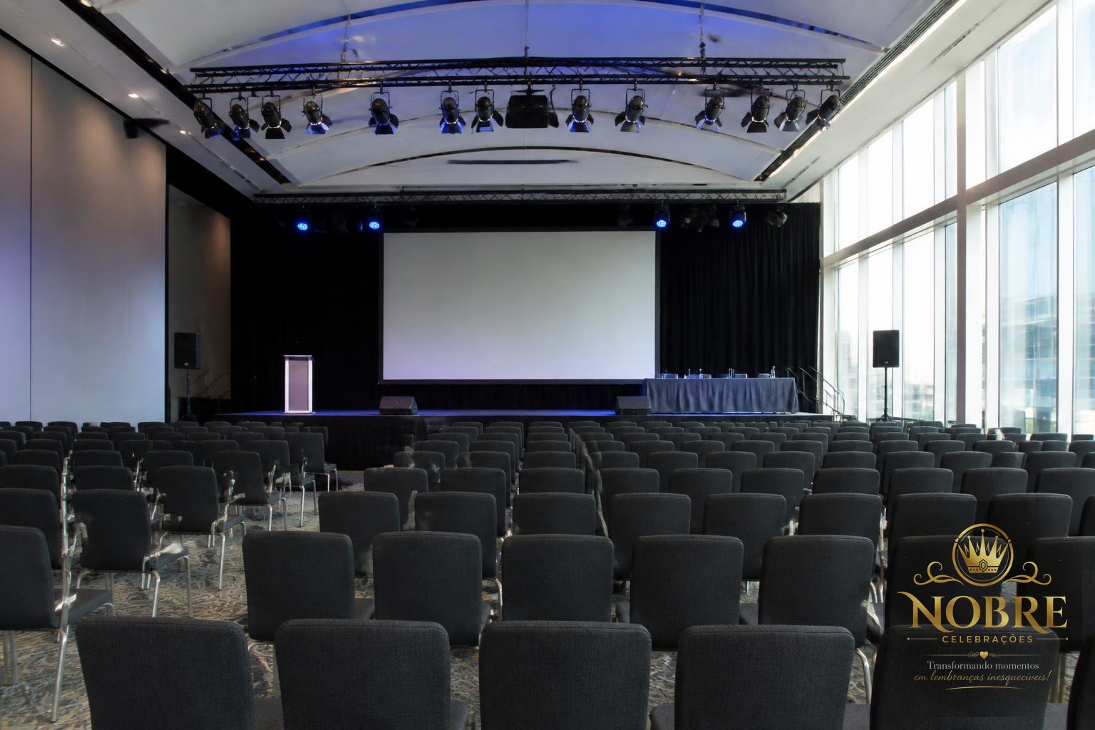 Ambiente preparado para conferência empresarial com palco, telão, iluminação profissional e cadeiras alinhadas