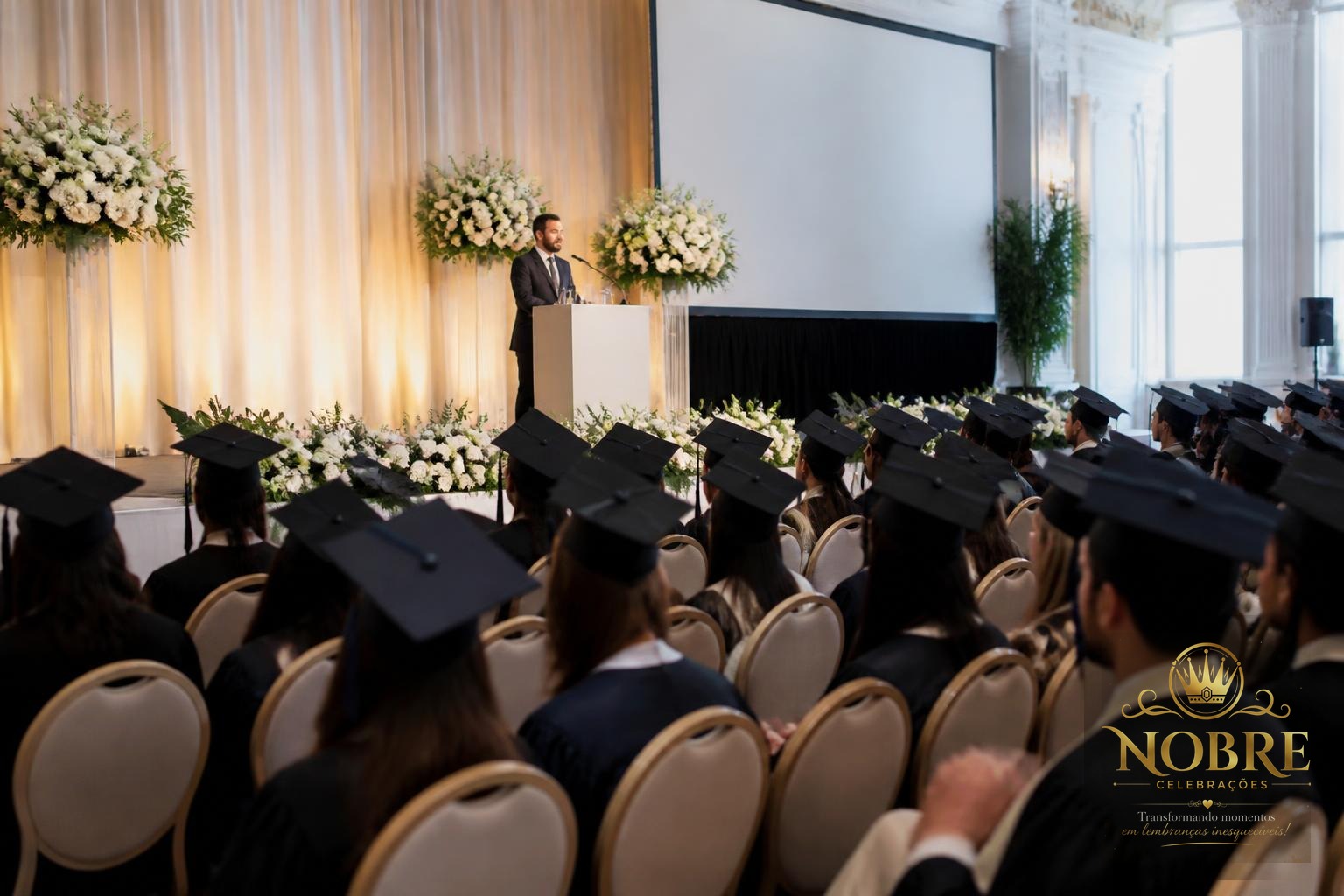 Cerimônia de formatura em salão amplo com palco elegante, púlpito, telão, iluminação profissional e arranjos de flores naturais brancas