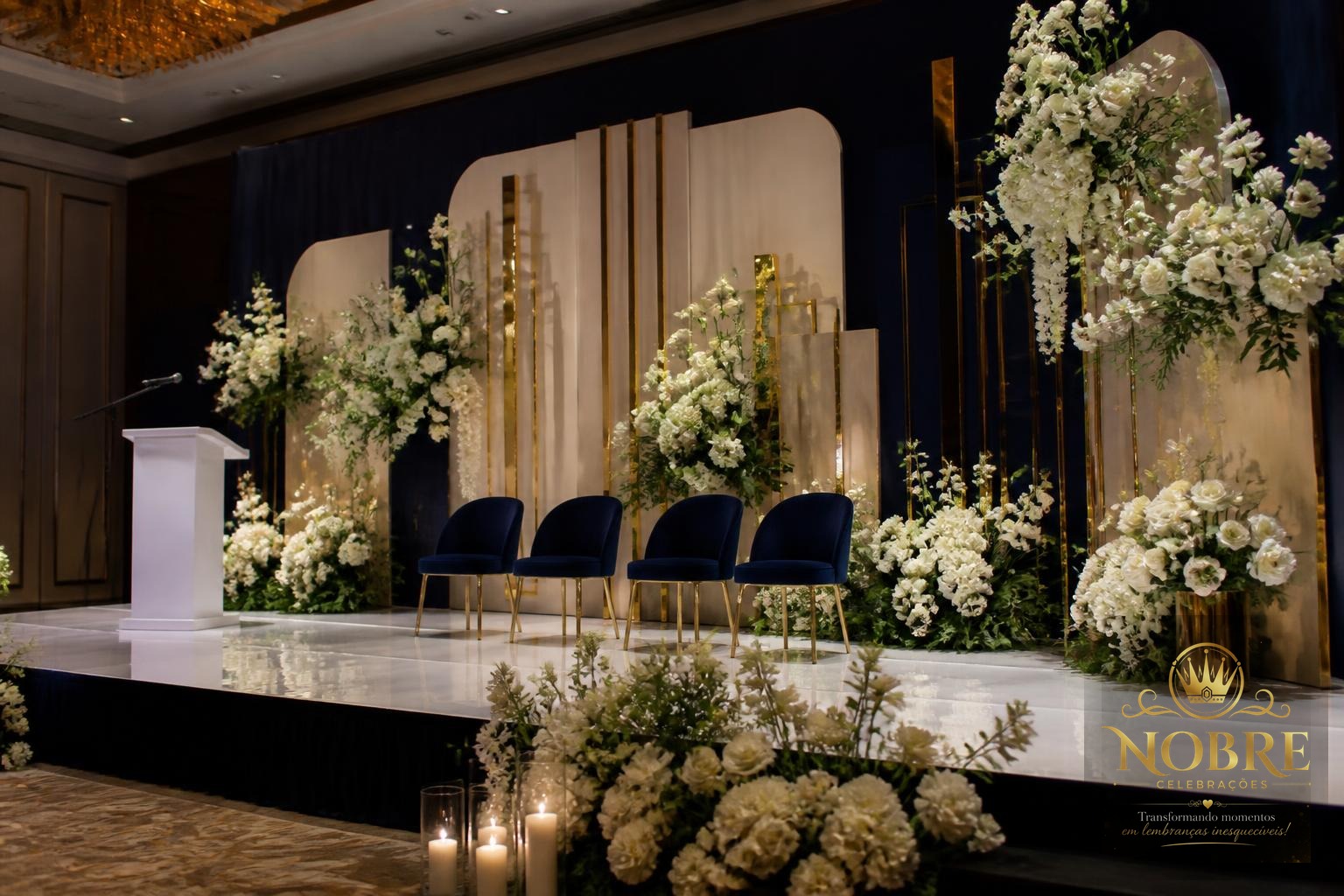 Palco principal de formatura com painel decorativo, iluminação cênica, flores naturais brancas e detalhes dourados discretos